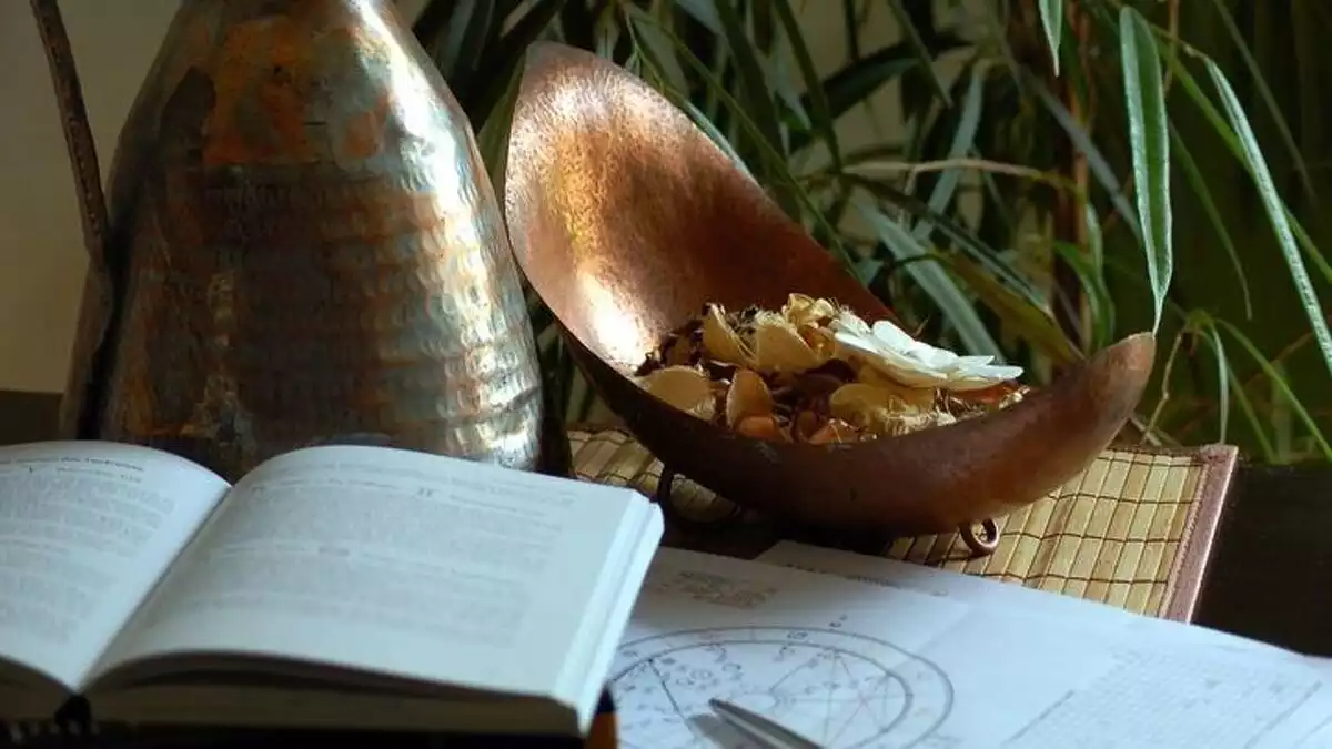Una mesa donde se puede ver un libro, un plano astrológico y un jarrón