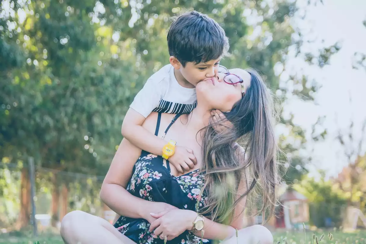 Imagen de una madre sentada en la hierba con tu hijo besándole por detrás