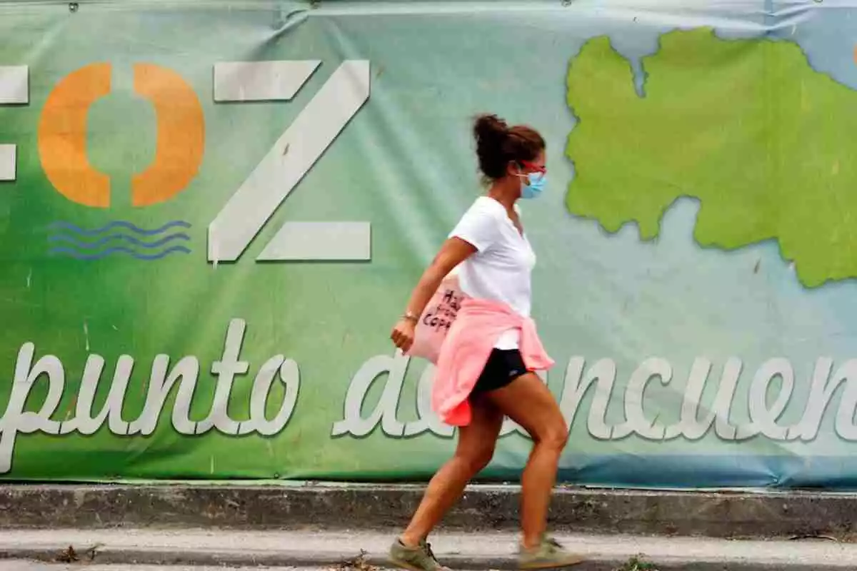 Una mujer con mascarilla camina frente a un cartel publicitario del pueblo de A Mariña en Lugo
