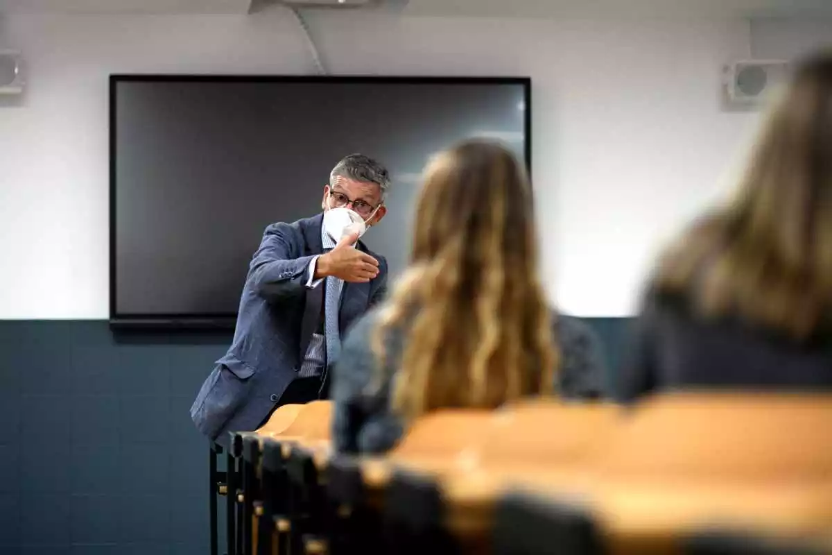 Un profesor dando una clase en una universidad con mascarilla y dos alumnas de espaldas
