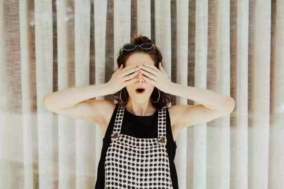 Una mujer vertida con una camiseta negra y un peto a cuadros se tapa los ojos mientras mantiene la boca abierta en señal de sorpresa