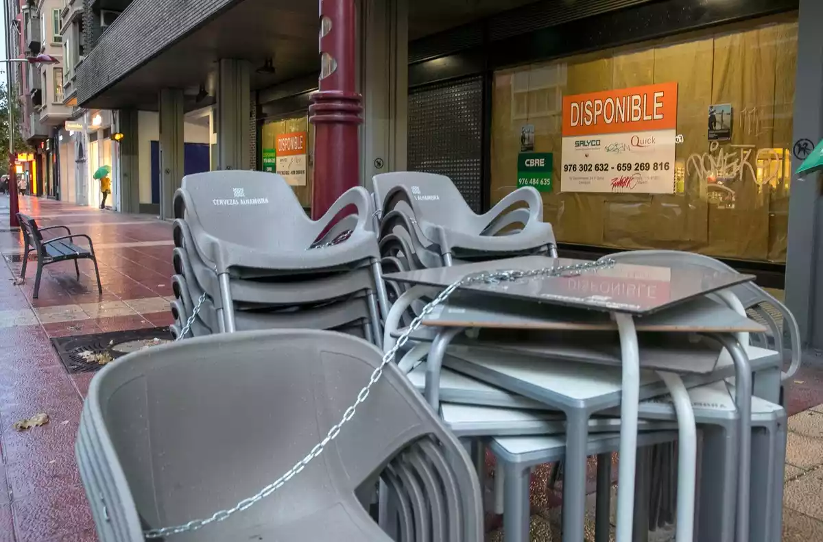 Imagen de mesas y sillas recogidas en la terraza de un bar con un cartel de alquiler de fondo