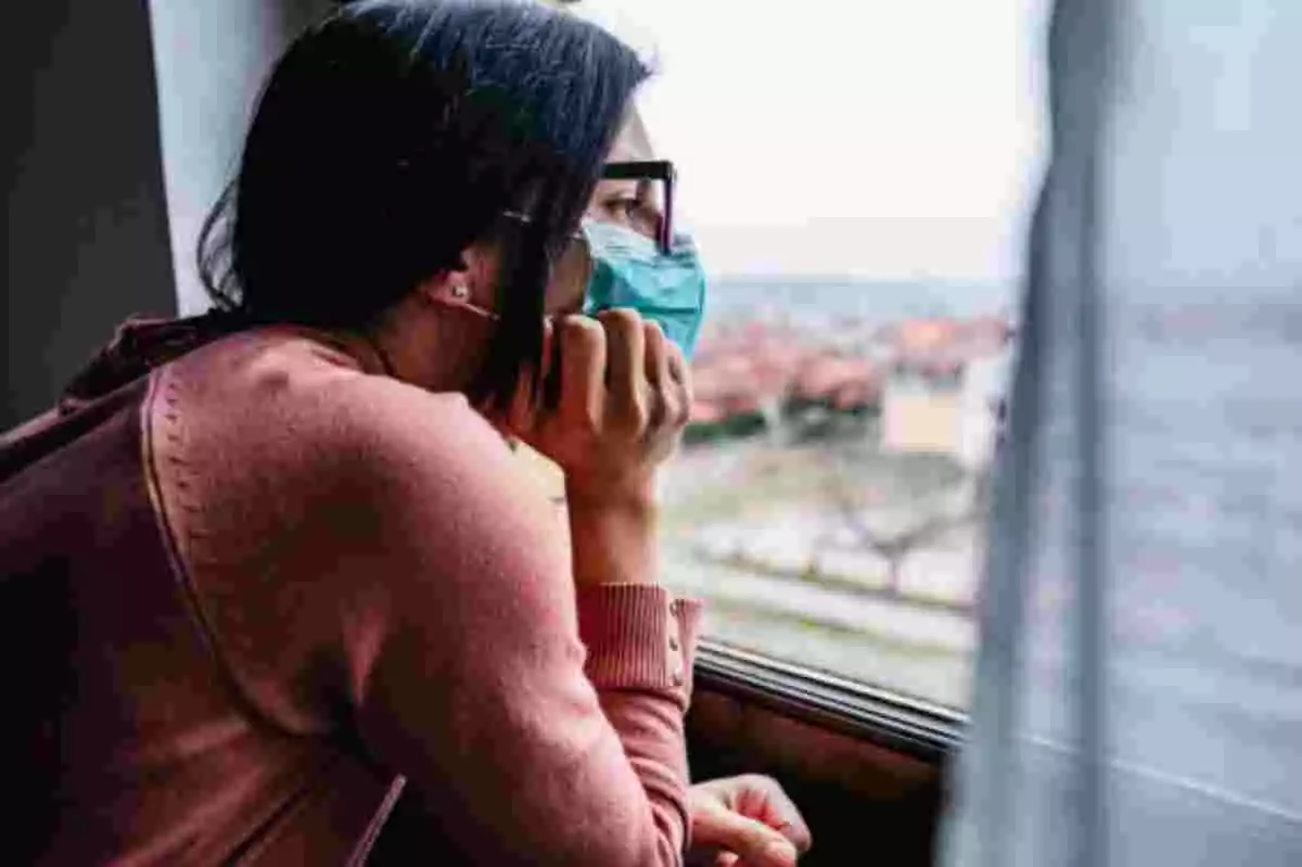Mujer con pose de aburrida y mascarilla mirando por la ventana