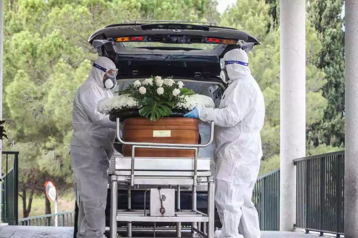 Dos personas con equipos de protección contra el Covid metiendo un féretro en un coche funebre