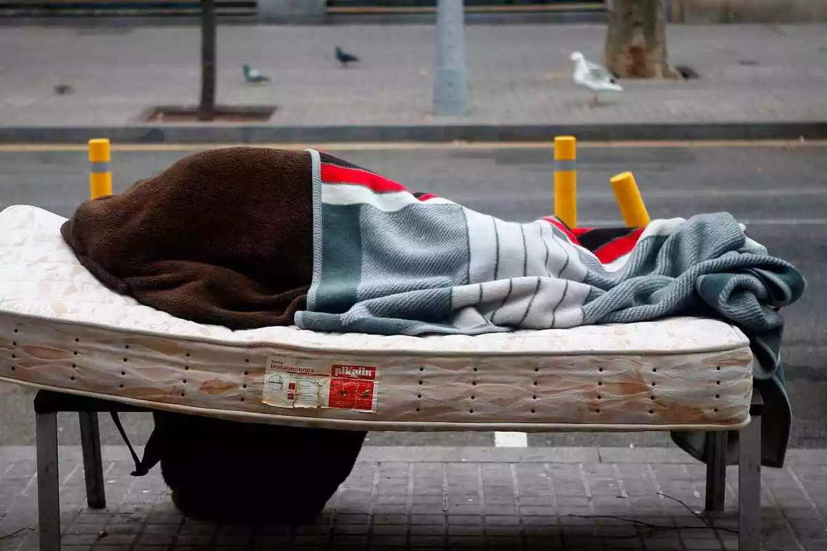 Una persona sin techo encima de un colchón, cubierto por una manta