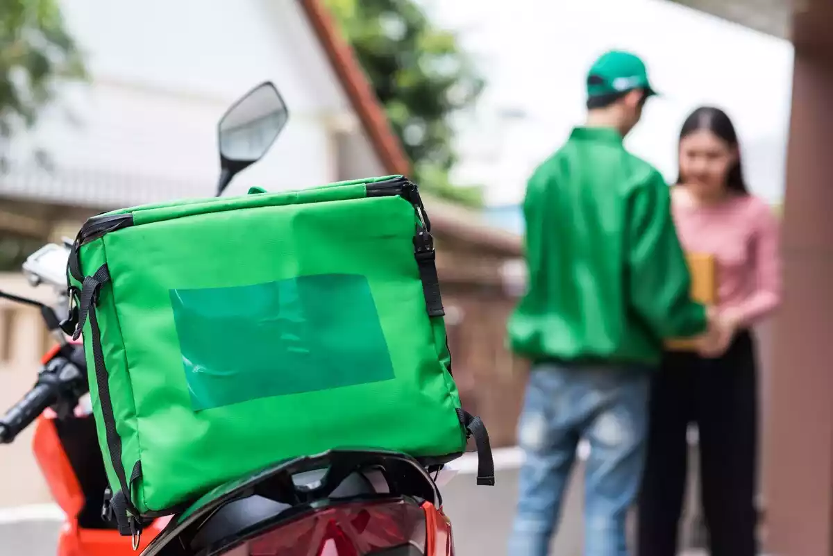 Un hombre entrega una bolsa de comida a domicilio a una clienta con la moto y la mochila en primer plano