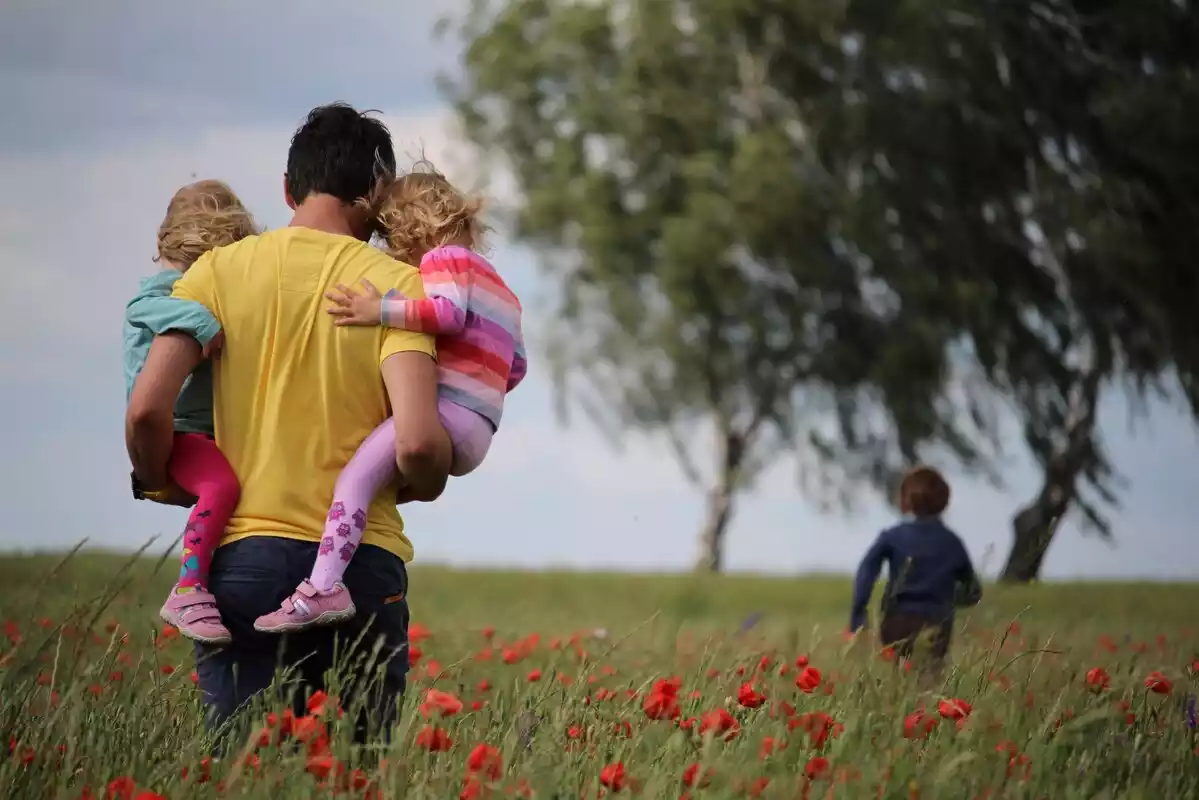 Imagen de archivo de un padre con dos niñas en brazos y un niño corriendo en el campo