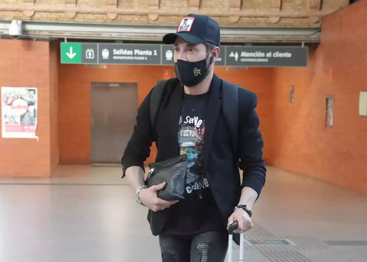 Plano medio de Antonio David con gorra y mascarilla y con una bolsa en la mano en una estación de tren