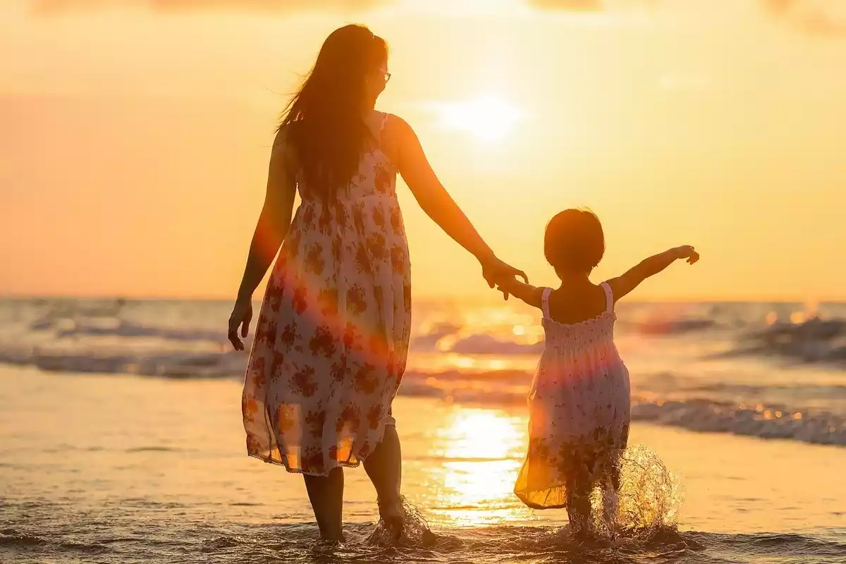 Una madre con su hija cogida de la mano en la playa con el sol por delante