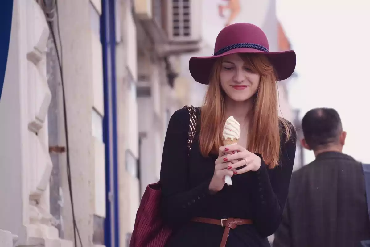 Una mujer joven con sobrero comiendo un helado de vainilla en la calle