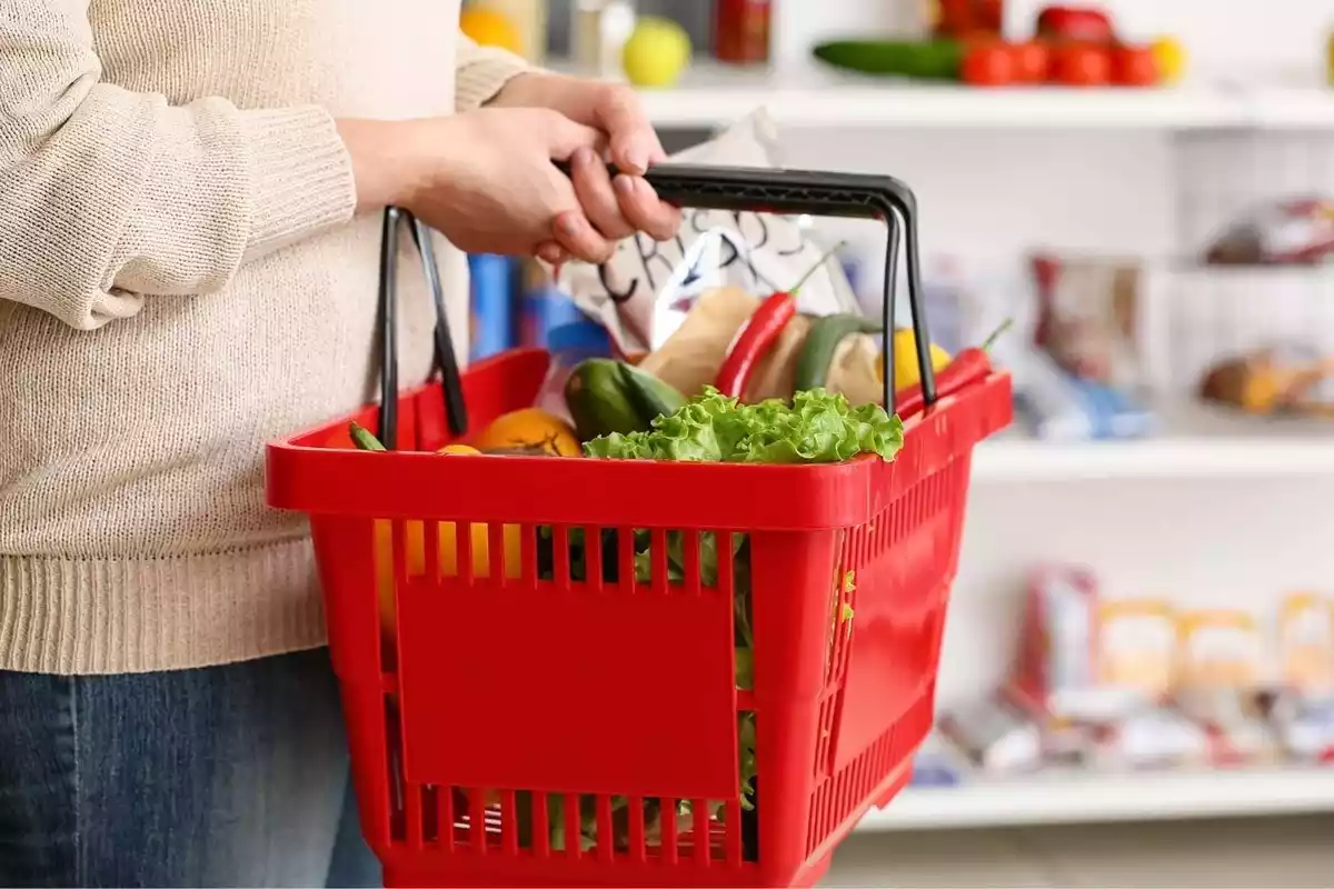 Persona en un supermercado cogiendo una cesta de la compra roja llena de verduras
