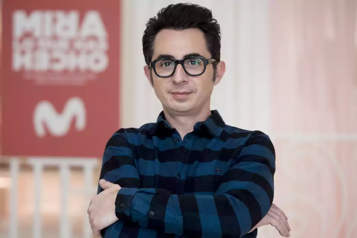Un hombre con gafas y camisa de cuadros posa con los brazos cruzados frente a un cartel rojo.