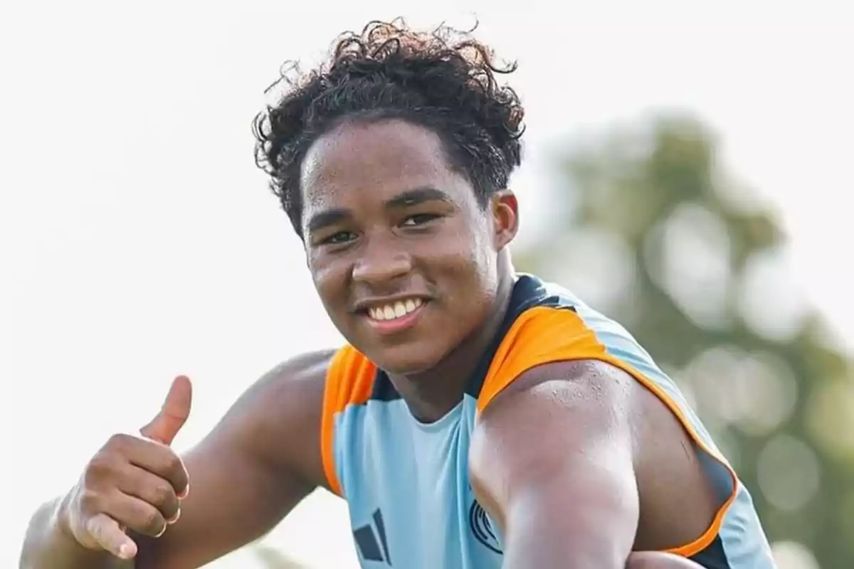Un joven deportista sonríe y hace un gesto de aprobación con el pulgar hacia arriba mientras lleva una camiseta sin mangas de color azul claro con detalles naranjas. Un joven deportista sonríe y hace un gesto de aprobación con el pulgar hacia arriba mientras lleva una camiseta sin mangas de color azul claro con detalles naranjas.