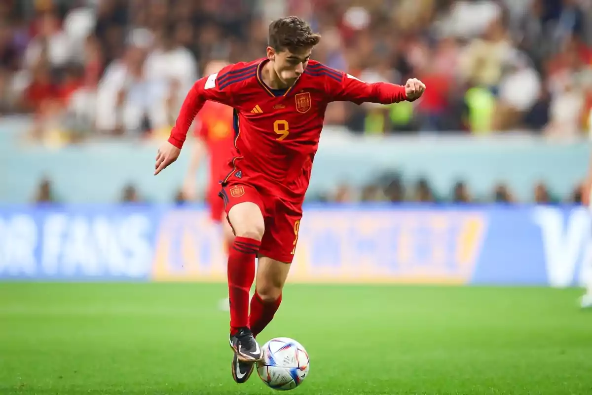 Futbolista de la selección de España con uniforme rojo controlando el balón durante un partido en un estadio lleno de espectadores Futbolista de la selección de España con uniforme rojo controlando el balón durante un partido en un estadio lleno de espectadores