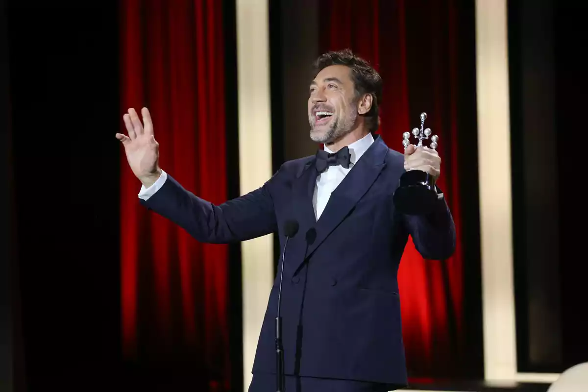 Javier Bardem en un escenario con un traje oscuro sosteniendo un trofeo y sonriendo mientras habla al micrófono. Javier Bardem en un escenario con un traje oscuro sosteniendo un trofeo y sonriendo mientras habla al micrófono.