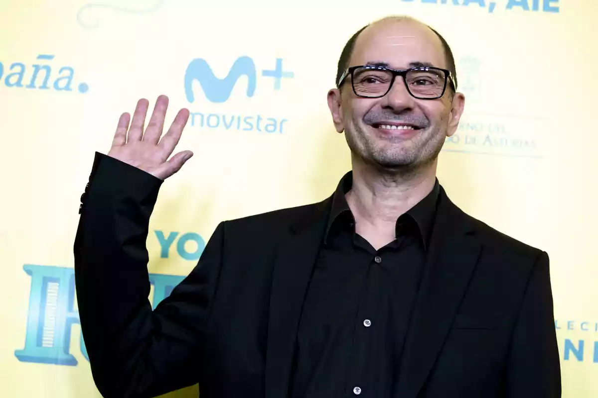 Un hombre con gafas y traje oscuro sonríe y saluda con la mano levantada frente a un fondo amarillo con logotipos.