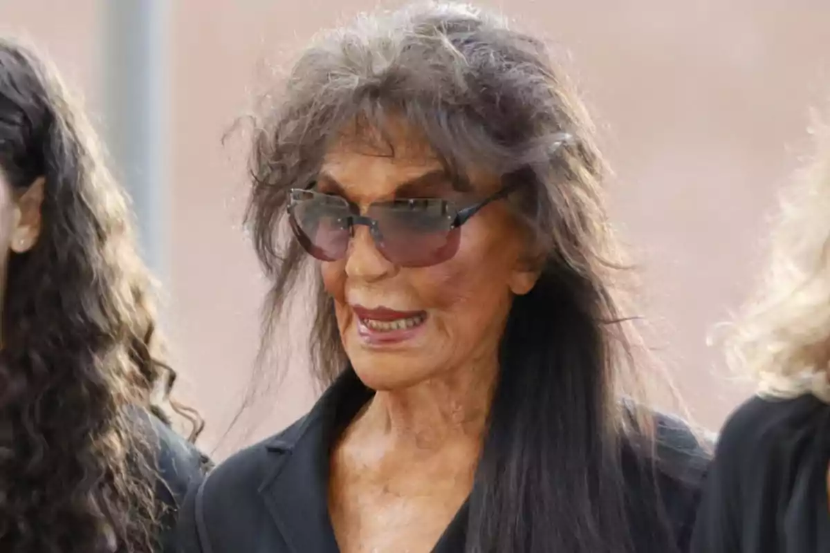 Una persona con gafas de sol y cabello largo y oscuro viste de negro. Una persona con gafas de sol y cabello largo y oscuro viste de negro.