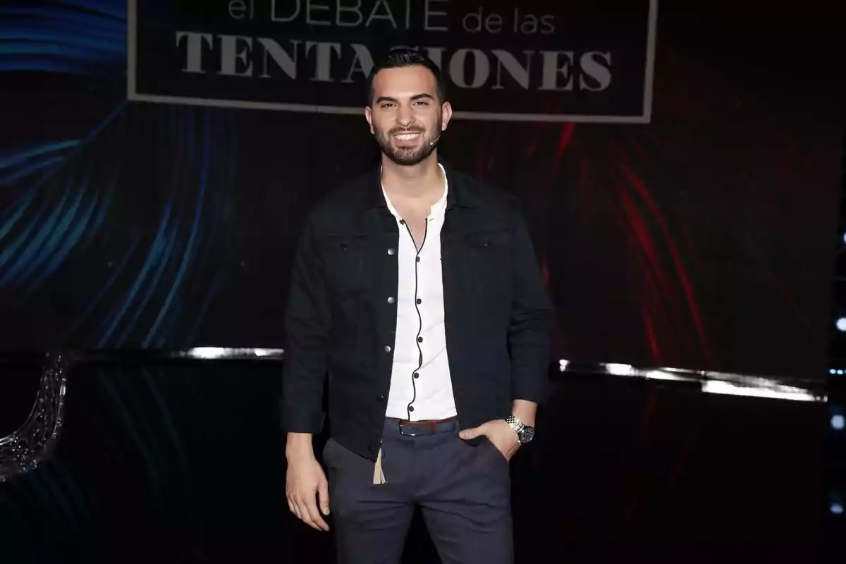 Un hombre sonriente con barba y cabello corto viste una chaqueta oscura y camisa blanca de pie frente a un fondo con luces azules y rojas.